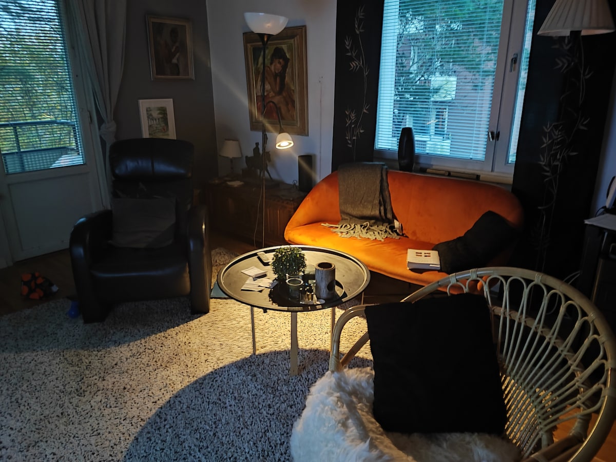 A cozy living area is presented, featuring a curved orange sofa beside a black armchair. A round table sits at the center, adorned with a plant and a mug. Warm light casts gentle shadows, and a textured rug covers the floor, enhancing the inviting setting.