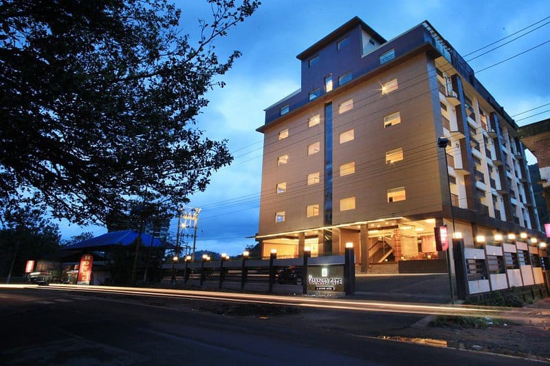 The exterior of the multi-story building is showcased at dusk, with bright windows illuminated in a warm glow. The entrance is marked by a well-lit sign, and trees are visible lining the street, creating a calm urban atmosphere.