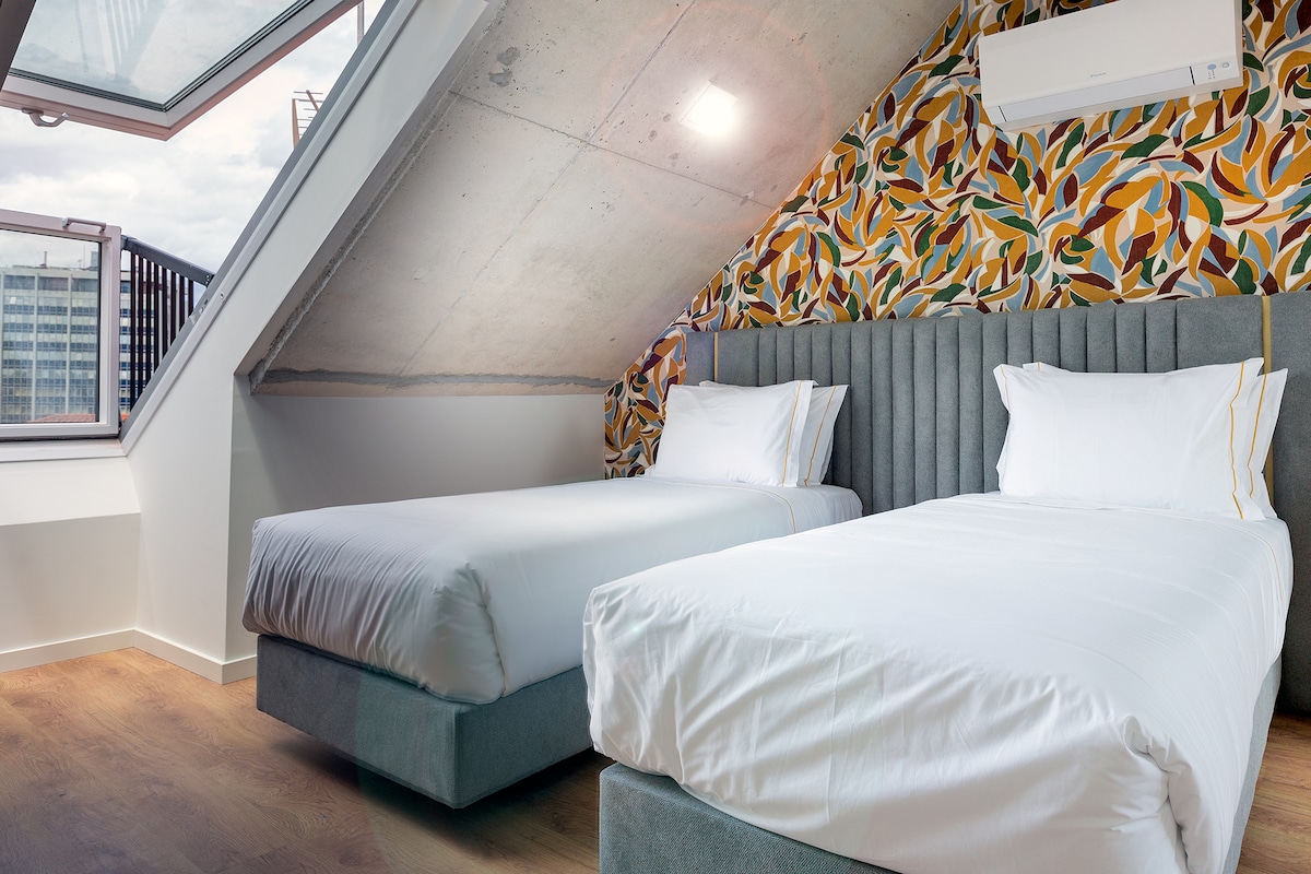 A bright bedroom area features two spacious beds with clean white linens, complemented by patterned wallpaper that adds a touch of colour. A large window offers natural light and city views, while an air conditioning unit is mounted on the ceiling for added comfort.