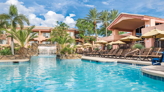 A resort-style swimming pool is surrounded by lounge chairs and umbrellas, creating a relaxed atmosphere. Lush palm trees are positioned along the pool area, complemented by a decorative waterfall feature. The building structure in the background follows a southwestern architectural style.