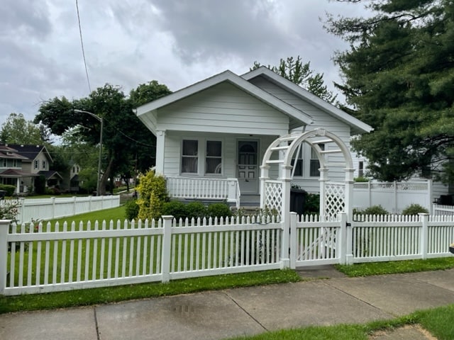 A charming home is set behind a white picket fence, surrounded by a well-maintained lawn. An inviting archway leads to the front steps. The single-story structure features multiple windows with white trim, allowing natural light into the living spaces.