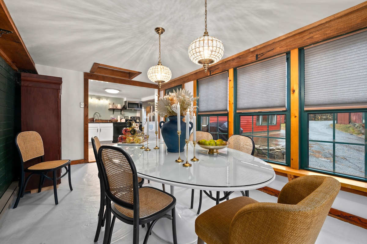 A bright dining room features a white oval table surrounded by six chairs, with large windows showcasing views of the outdoors. Two pendant lights hang from the ceiling, and a decorative vase with flowers sits center stage, complemented by fruit bowls on the table.