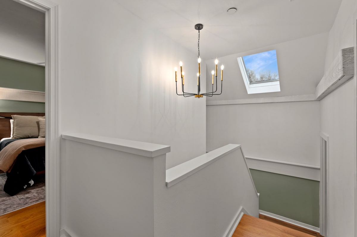 A well-lit staircase area showcases a modern chandelier above, casting a warm glow. The walls are painted in crisp white, while a skylight allows natural light to brighten the space. Soft green accents provide a subtle contrast, enhancing the airy feel of the upper level.