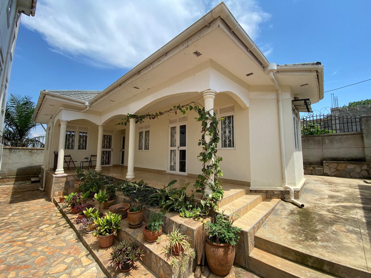 The exterior of the cottage showcases a spacious porch supported by decorative columns, with potted plants lining the steps. A smooth stone pathway leads towards the entrance, under a clear blue sky, creating a serene and inviting atmosphere.