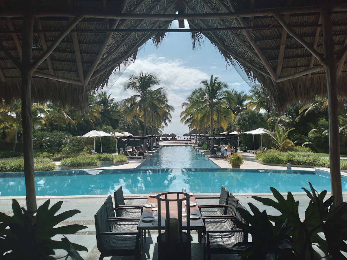 A spacious resort pool area is framed by a thatched-roof gazebo. Clear blue waters reflect palm trees and sun loungers under umbrellas. Lush greenery surrounds the pool, inviting relaxation and enjoyment of the tropical environment.