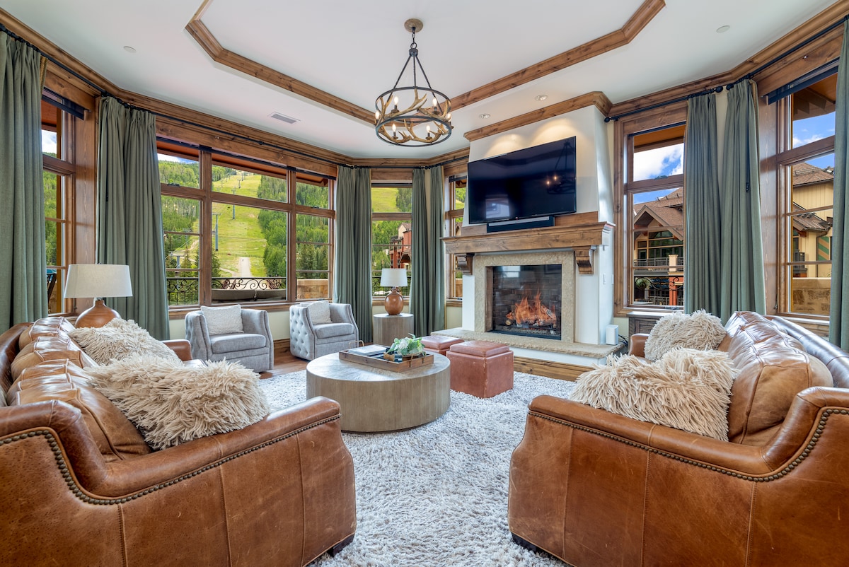 A spacious living area features large windows that offer views of the surrounding nature. Comfortable leather sofas are arranged around a round coffee table, with a cozy area rug beneath. A television is mounted above a decorative fireplace, enhancing the inviting atmosphere.