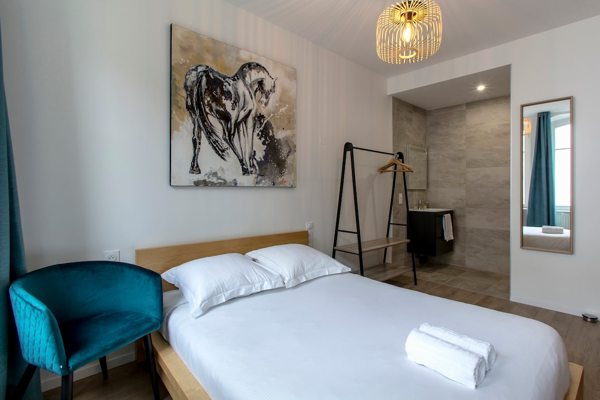 A well-lit bedroom features a spacious bed with white linens. An artistic horse painting adorns the wall above the bed. A compact corner workspace is visible, along with a full-length mirror and a clothing rack. Natural light enters through large windows draped with teal curtains.