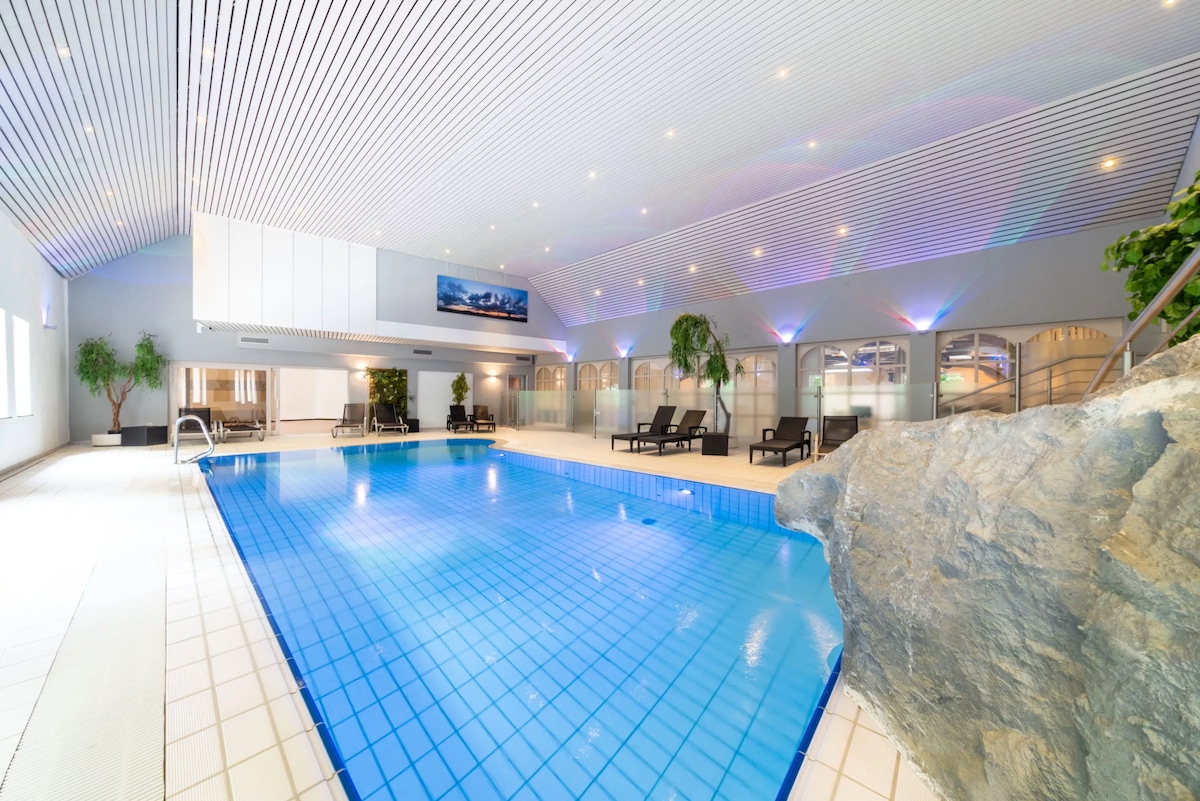 An indoor swimming pool is surrounded by a sleek, light-filled space. The pool is flanked by lounge chairs and features natural rock accents. Soft lighting enhances the contemporary design, creating a calm atmosphere, suitable for relaxation and enjoyment.