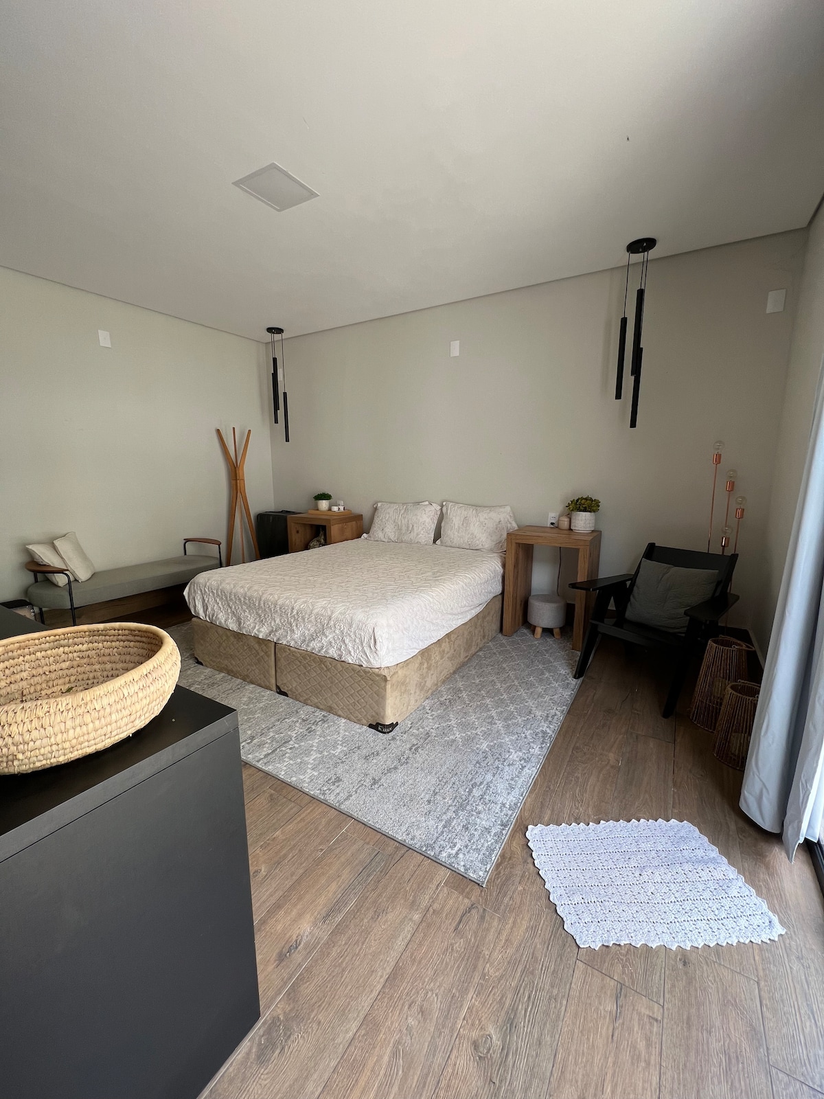 A neatly arranged bedroom features a spacious bed with light-colored bedding. Two bedside tables with plants are positioned beside the bed. A textured area rug lies beneath the bed, complementing the wooden flooring. A chair and a simple coat rack are visible in the corner.