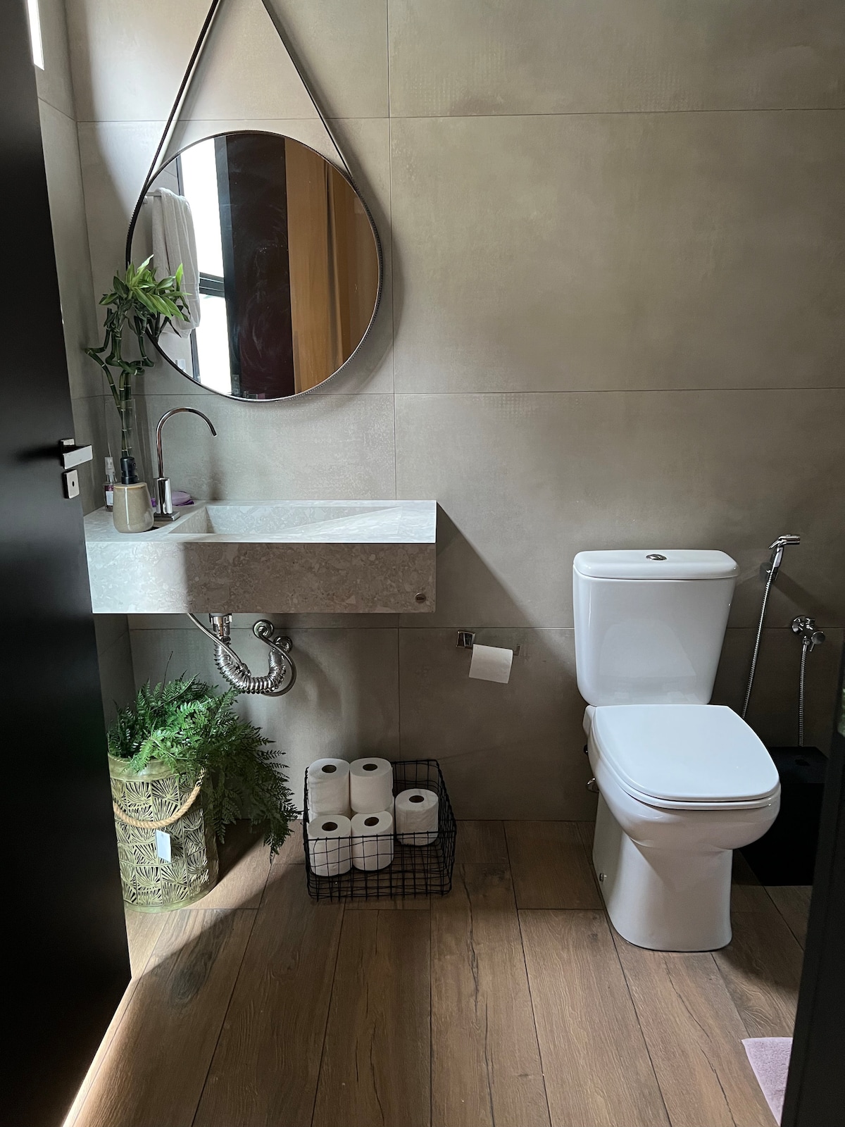A modern bathroom features a minimalist sink with a wall-mounted mirror. Fresh greenery is positioned next to the sleek toilet, while a wire basket holds multiple rolls of toilet paper on the wooden floor.