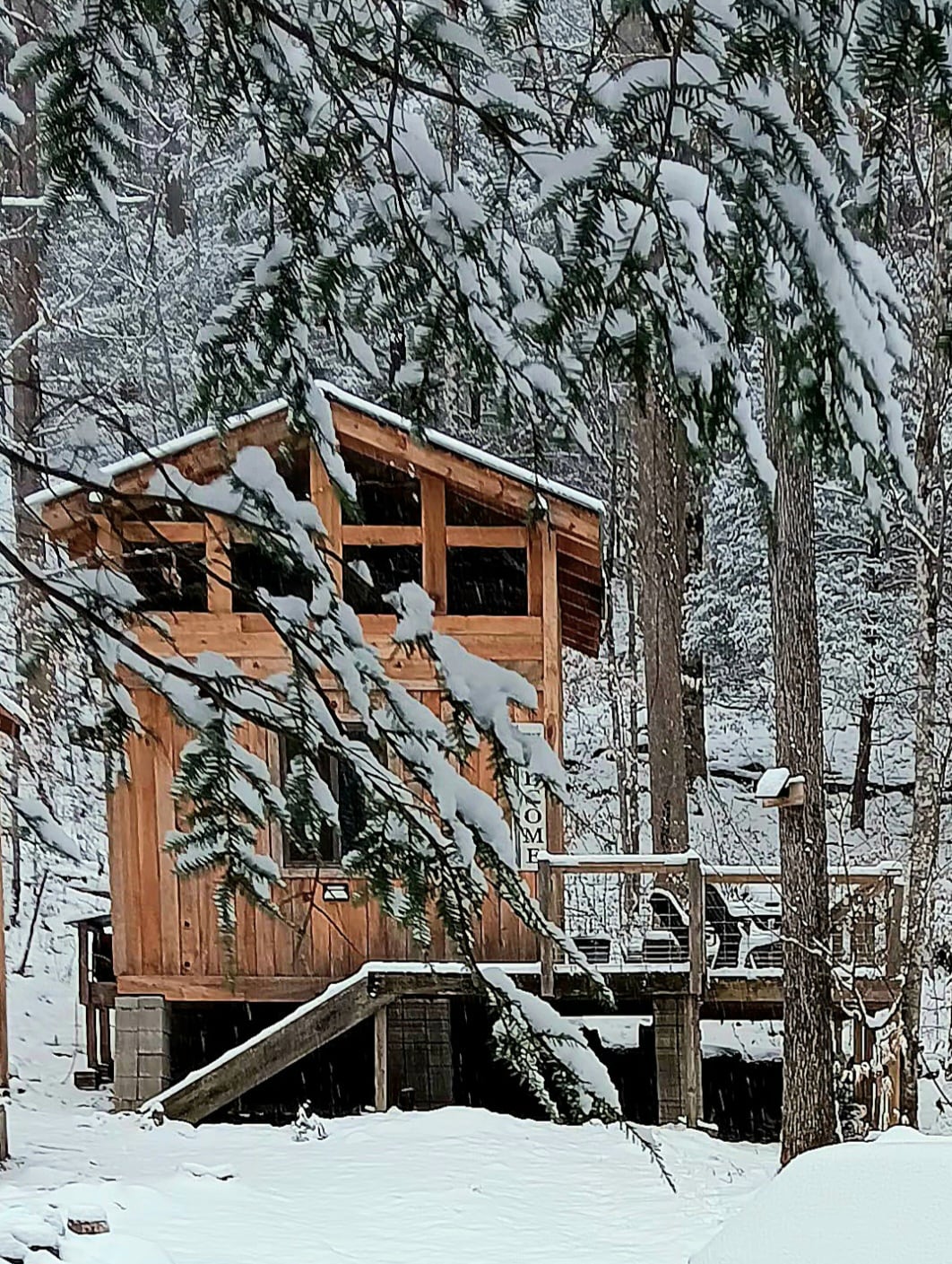 A rustic wooden cabin is framed by snow-covered trees, with a deck featuring railings and steps leading up to the entrance. Snow blankets the ground, creating a serene winter landscape around the structure.