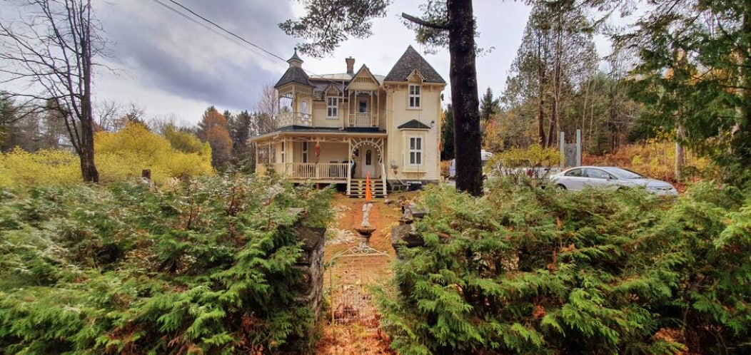A historic building is framed by lush greenery, showcasing a charming two-story structure with intricate architectural details. The porch is inviting, featuring a staircase leading to the entrance. The surrounding area is dotted with trees, offering a serene atmosphere.
