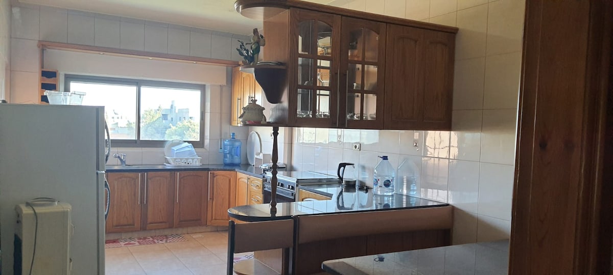 The kitchen features wooden cabinetry and a large window that allows natural light to fill the space. Appliances are visible, including a stove and a refrigerator. A countertop with bar seating is present, enhancing the kitchen's spacious feel.