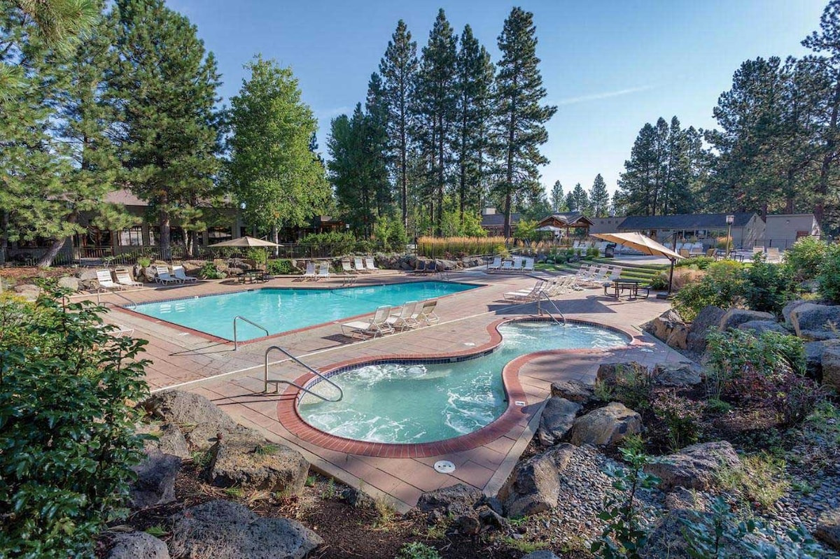 A well-maintained outdoor pool area is surrounded by lush trees and decorative rocks. The larger pool is complemented by a smaller hot tub, with ample lounge chairs positioned around the water for relaxation. Sunlight brightens the scene, highlighting the natural landscape.