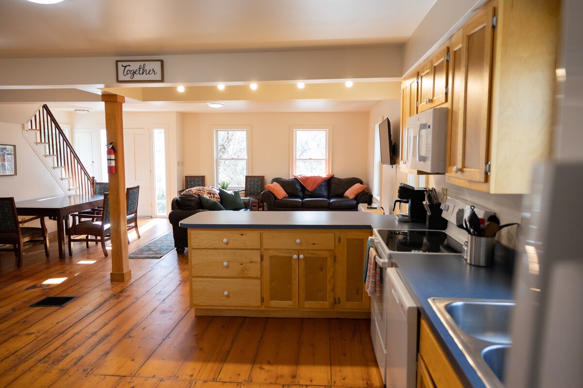The open concept living area showcases a full kitchen with wooden cabinetry and a breakfast bar. A dining table for six to eight people is visible. The living room area features a couch, TV, and windows that allow natural light to brighten the space.