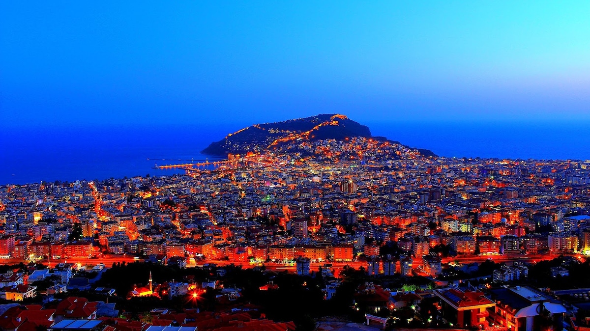 A panoramic view of a coastal city at twilight, showcasing a vibrant array of lights across the landscape. The illuminated hillside merges with the darkening sky, while the water reflects the glowing lights, creating a peaceful night scene.