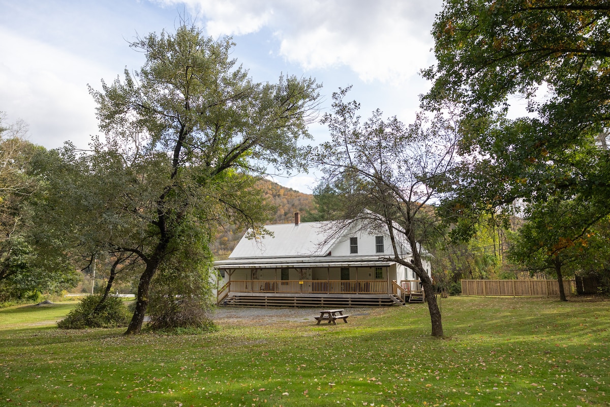 The exterior of a charming country home is framed by mature trees and a spacious lawn. A covered porch extends along the front, offering seating options. A picnic table is positioned on the grass, inviting outdoor gatherings amidst a peaceful natural setting.