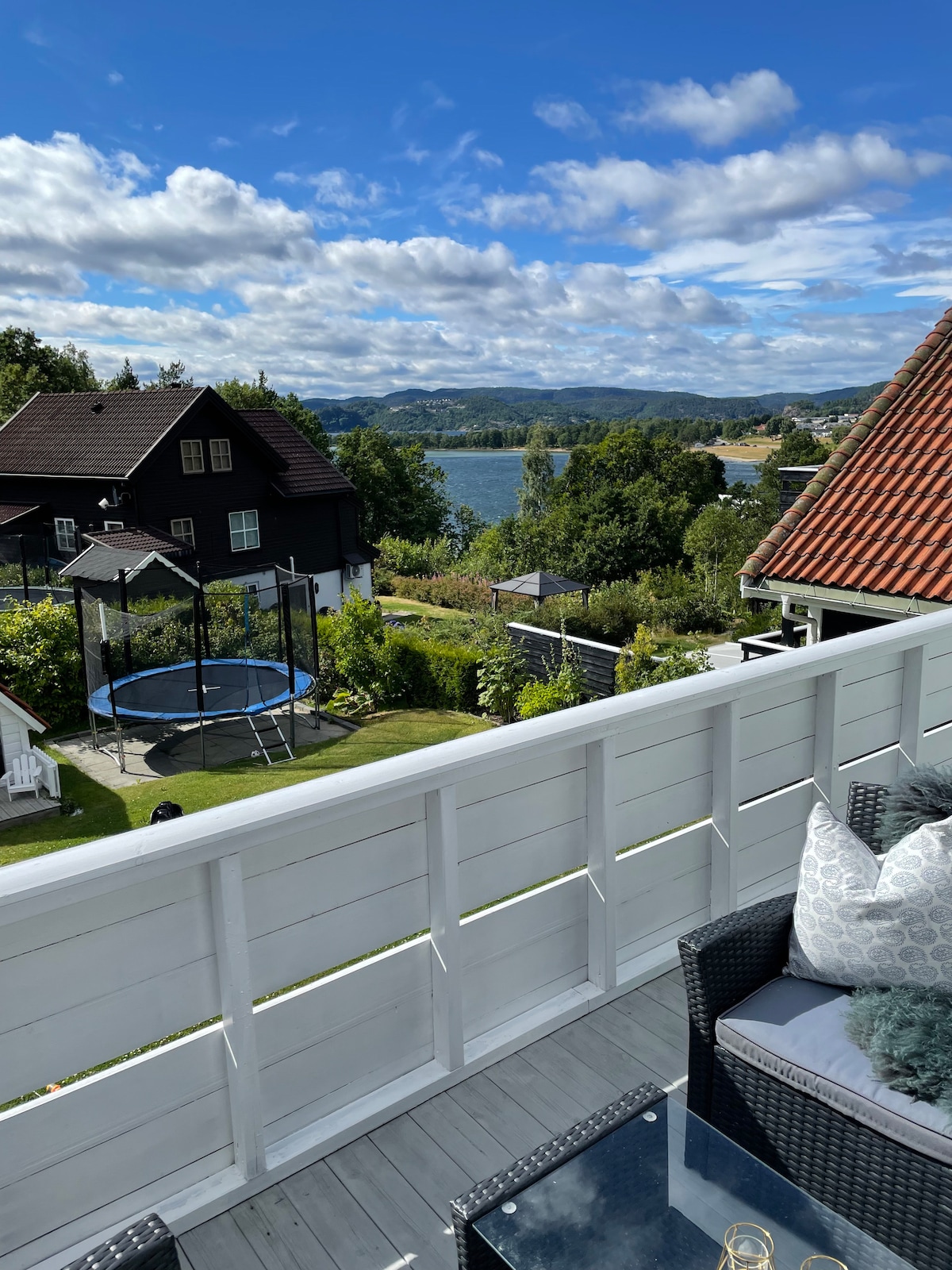 A balcony view captures a landscape with a trampoline in a lush garden, a shimmering body of water, and rolling hills in the distance. Comfortable seating with decorative cushions is positioned nearby, providing a space to relax and enjoy the scenery.