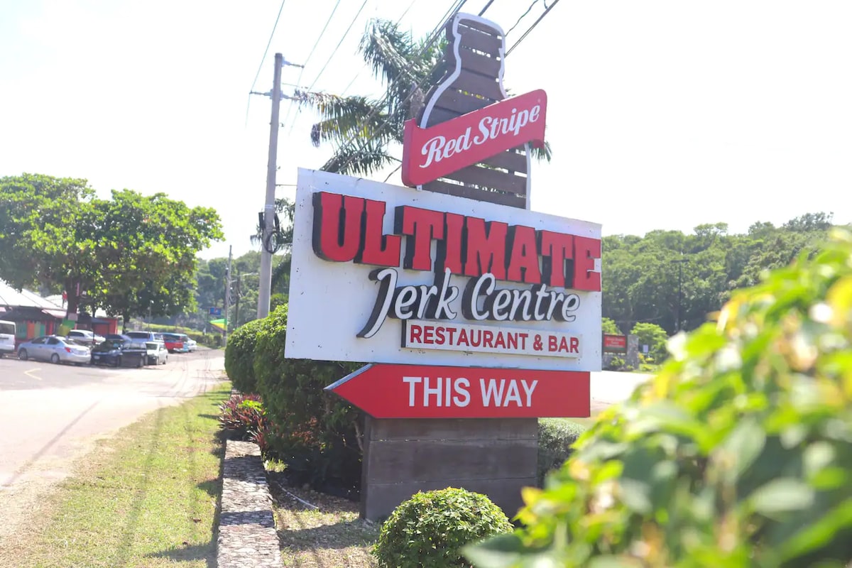 The entrance to Ultimate Jerk Centre is marked by a large, vibrant sign featuring the name prominently displayed. Surrounding greenery provides a natural frame, while the clear sky above enhances visibility. Traffic can be seen in the background, indicating accessibility to the restaurant and bar.