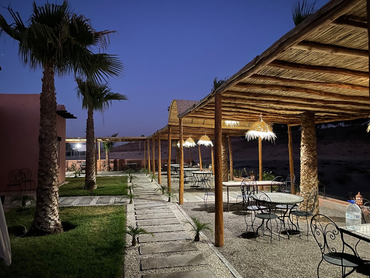 A well-lit outdoor seating area features several round tables with wrought iron chairs, shaded by a thatched roof. Palm trees and planted greenery enhance the inviting atmosphere, while stone pathways guide guests through the tranquil space.