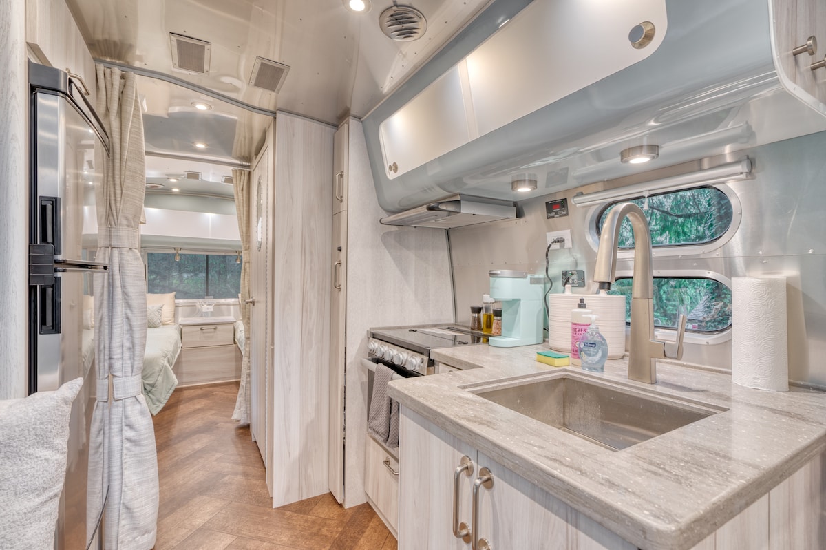 The Airstream's interior features a modern kitchen area with light-colored cabinetry and a marble-like countertop. A stainless steel sink and stove are visible, complemented by a color-coordinated blue appliance. Natural light enters through the windows, highlighting the warm wood flooring.