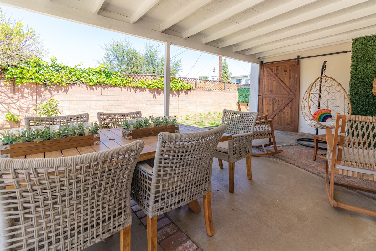 A shaded outdoor space features a rectangular wooden dining table surrounded by six woven chairs. A hanging chair is positioned nearby, and climbing plants adorn the walls. The concrete flooring enhances the open and spacious feel of this area.