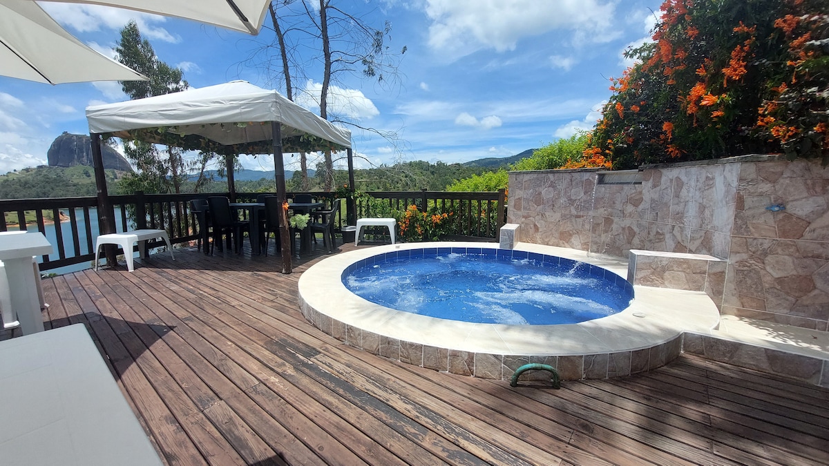 An outdoor area features a circular jacuzzi surrounded by a wooden deck, with white lounge chairs nearby. A shaded dining area is seen, complete with a table and chairs, all set against a backdrop of lush greenery and colorful flowers.