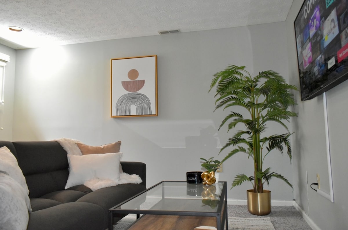 A cozy living area is highlighted by a dark sectional sofa adorned with light cushions. A glass coffee table sits in front, complemented by a small decorative piece. A tall potted plant adds a touch of greenery, while a wall-mounted television is visible alongside an abstract art piece.