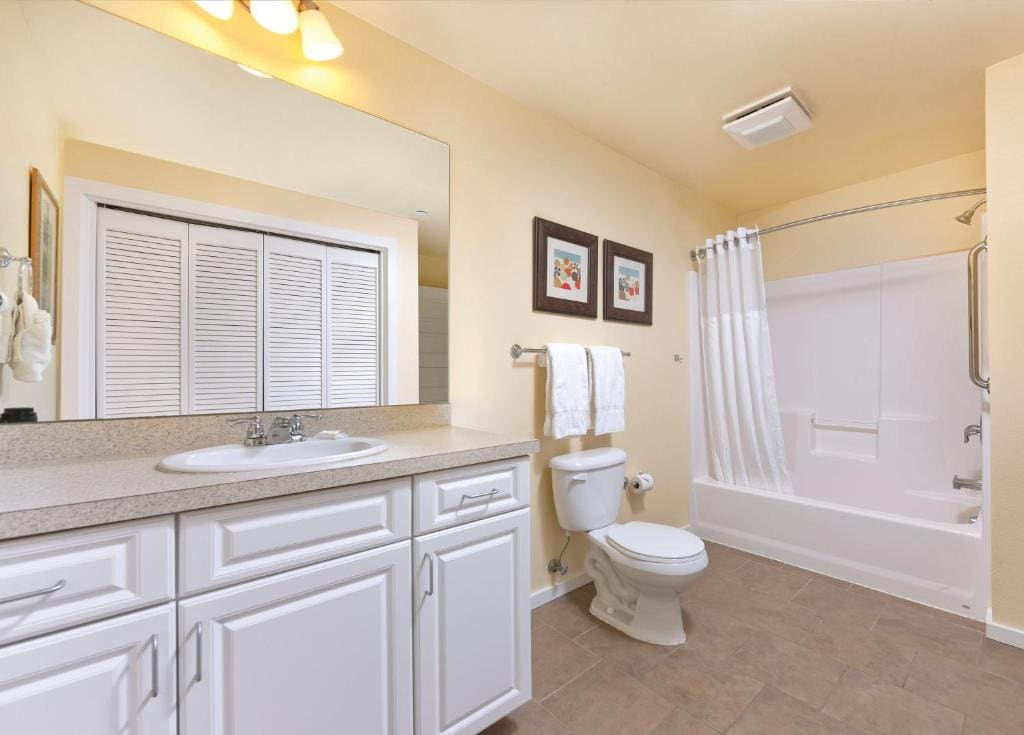 A well-lit bathroom features a spacious vanity with dual sinks and a large mirror above. The area includes a shower-tub combination with a white curtain, and towels are neatly hung. Neutral-colored walls and tiled flooring enhance the clean and organized space.