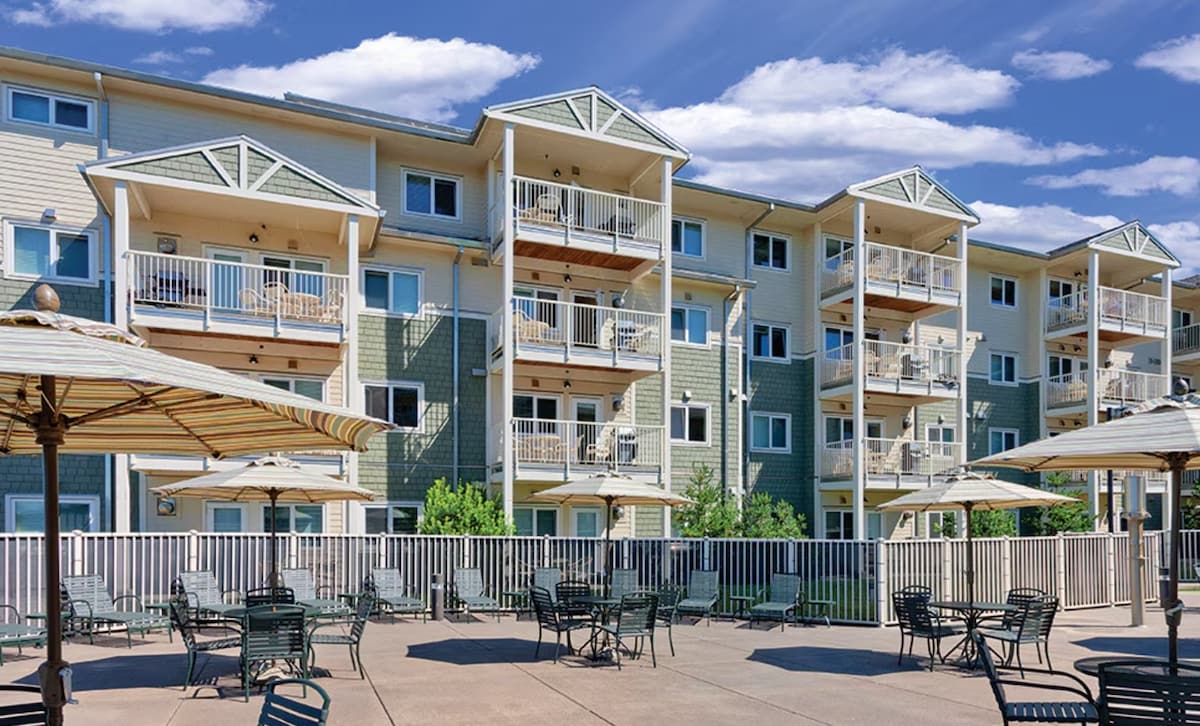 A multi-story building is presented, featuring balconies and ample windows. Patio seating with tables and chairs is visible in the foreground, surrounded by a low railing. Cloudy blue skies enhance the overall atmosphere of the outdoor space.