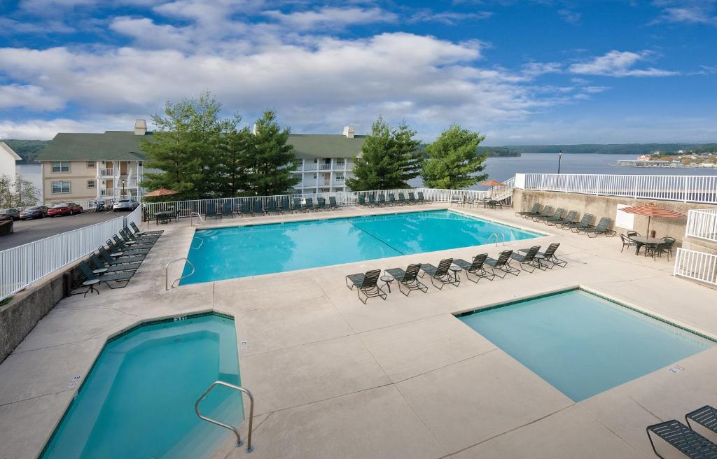 An outdoor pool area is showcased, featuring a large swimming pool surrounded by multiple lounge chairs. Two hot tubs are visible, one adjacent to the main pool. Lush greenery is present in the background, alongside views of the water under a partly cloudy sky.