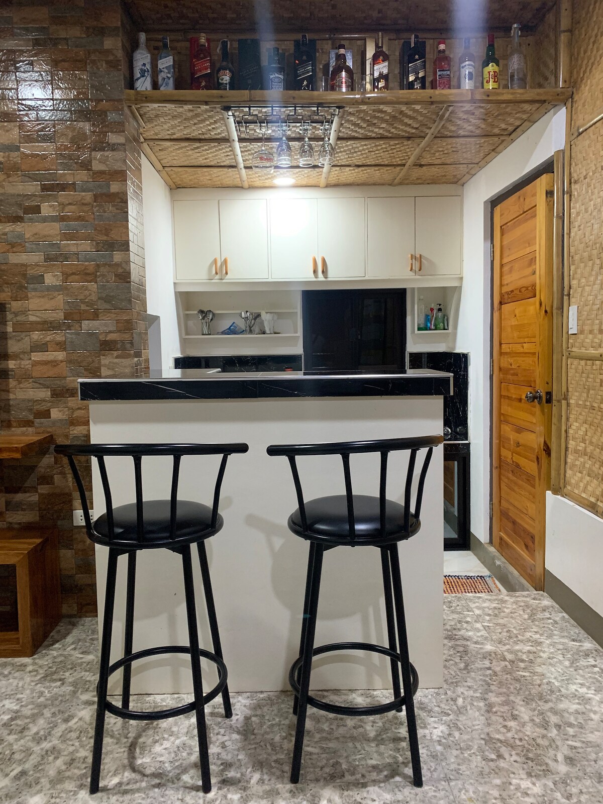 A compact kitchen area features a bar with two black stools. Above, a variety of bottles is displayed on a shelf. The kitchen cabinets are white, complemented by dark countertops. A refrigerator and appliances are visible, providing essential amenities for self-catering.