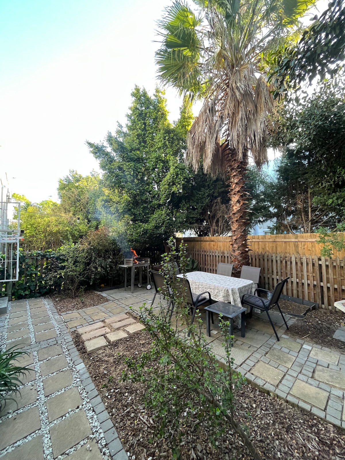 An outdoor braai area features a dining table surrounded by four chairs, set among pavers and garden beds. Lush green foliage provides a natural backdrop, while a palm tree adds height. Mild smoke rises from the grill, suggesting recent use.