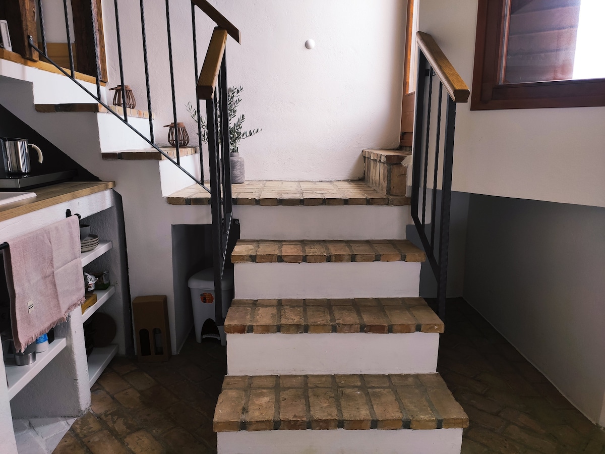 A set of tiled stairs leads up to a bright space, flanked by white walls and wooden accents. A small potted plant sits at the top, with a wooden door visible at the entrance. Kitchen essentials can be seen on shelving to the left.