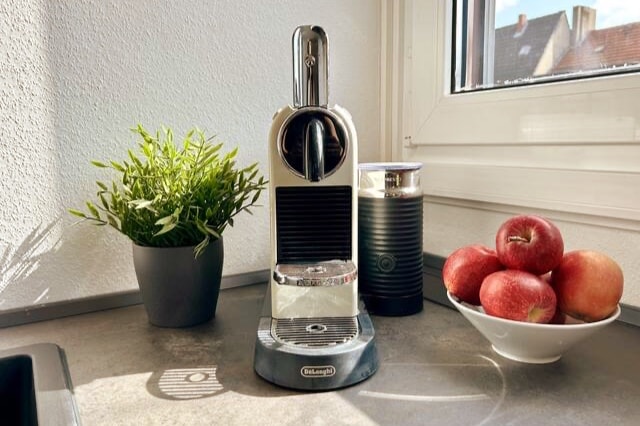 A sleek coffee machine is positioned on a countertop, accompanied by a small potted plant and a bowl of red apples. Natural light streams in through the window, highlighting the sturdy design of the machine and the fresh produce beside it.