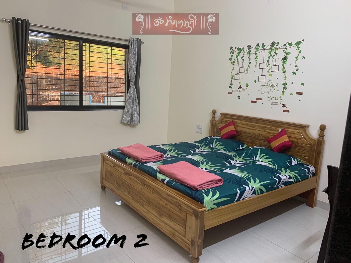 A bright bedroom features a spacious wooden bed adorned with tropical-patterned bedding. Soft towels are arranged neatly on the bed. Large windows allow natural light to fill the room while offering views of the surroundings. Simple wall decor adds a touch of charm.