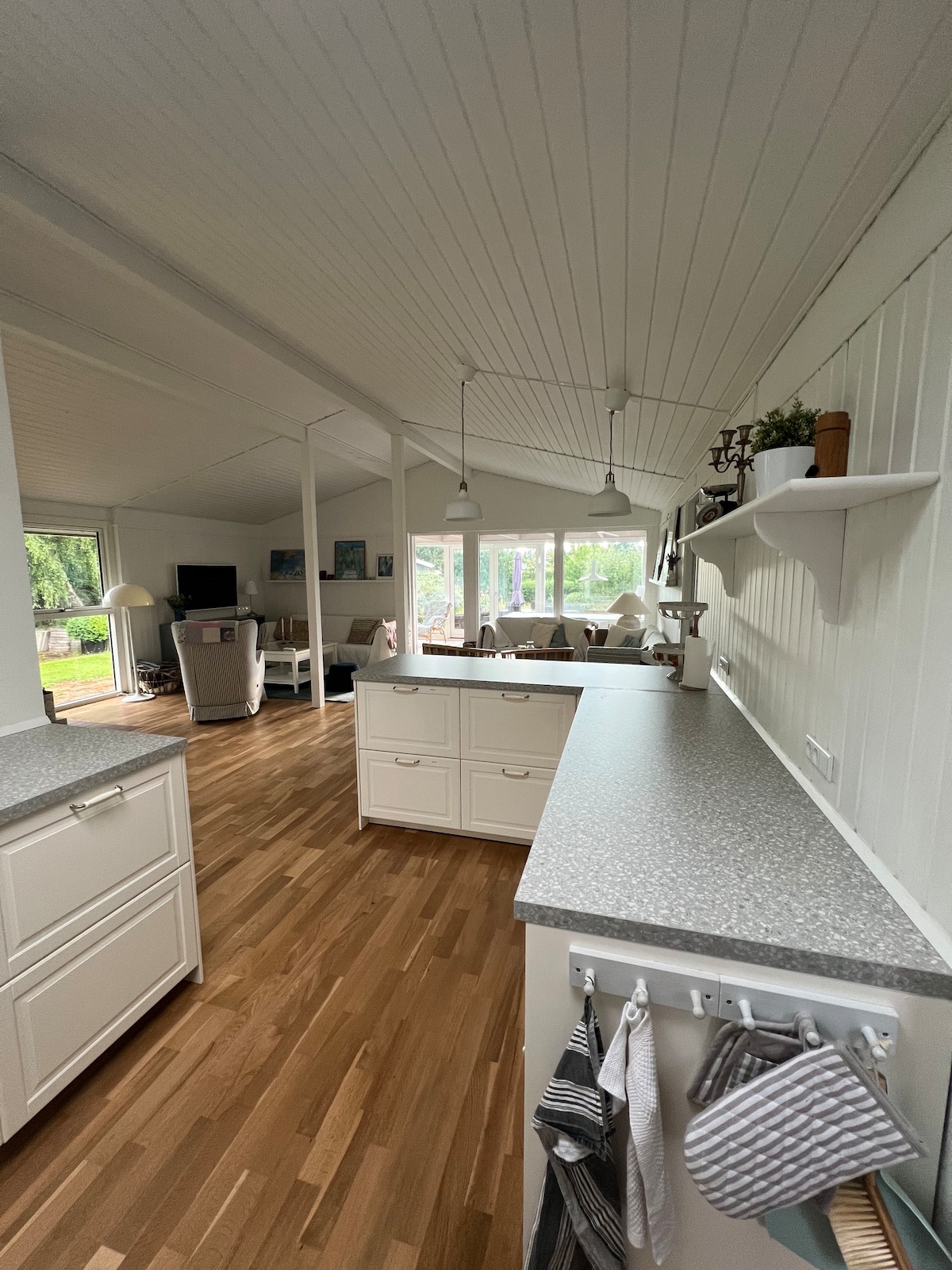 A well-lit kitchen area features a light-colored countertop, complemented by white cabinetry. Two pendant lights hang above the counter, while large windows bring in ample natural light. The open layout connects seamlessly to a cozy living area, enhancing the spaciousness of the home.