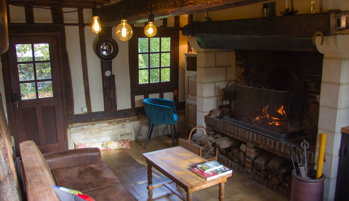 A traditional living area features a cozy sofa facing a stone fireplace, where flames are gently glowing. Warm lighting is provided by hanging bulbs, and a rustic wooden table occupies the center. A blue armchair adds a pop of color, complementing the space's inviting design.
