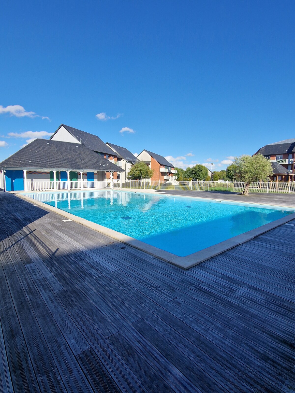 A spacious outdoor pool is surrounded by a wooden deck. Sunlight reflects off the clear water, inviting relaxation. Nearby buildings are visible against a bright sky, contributing to a serene environment. The area is designed for quiet enjoyment during the pool's seasonal hours.