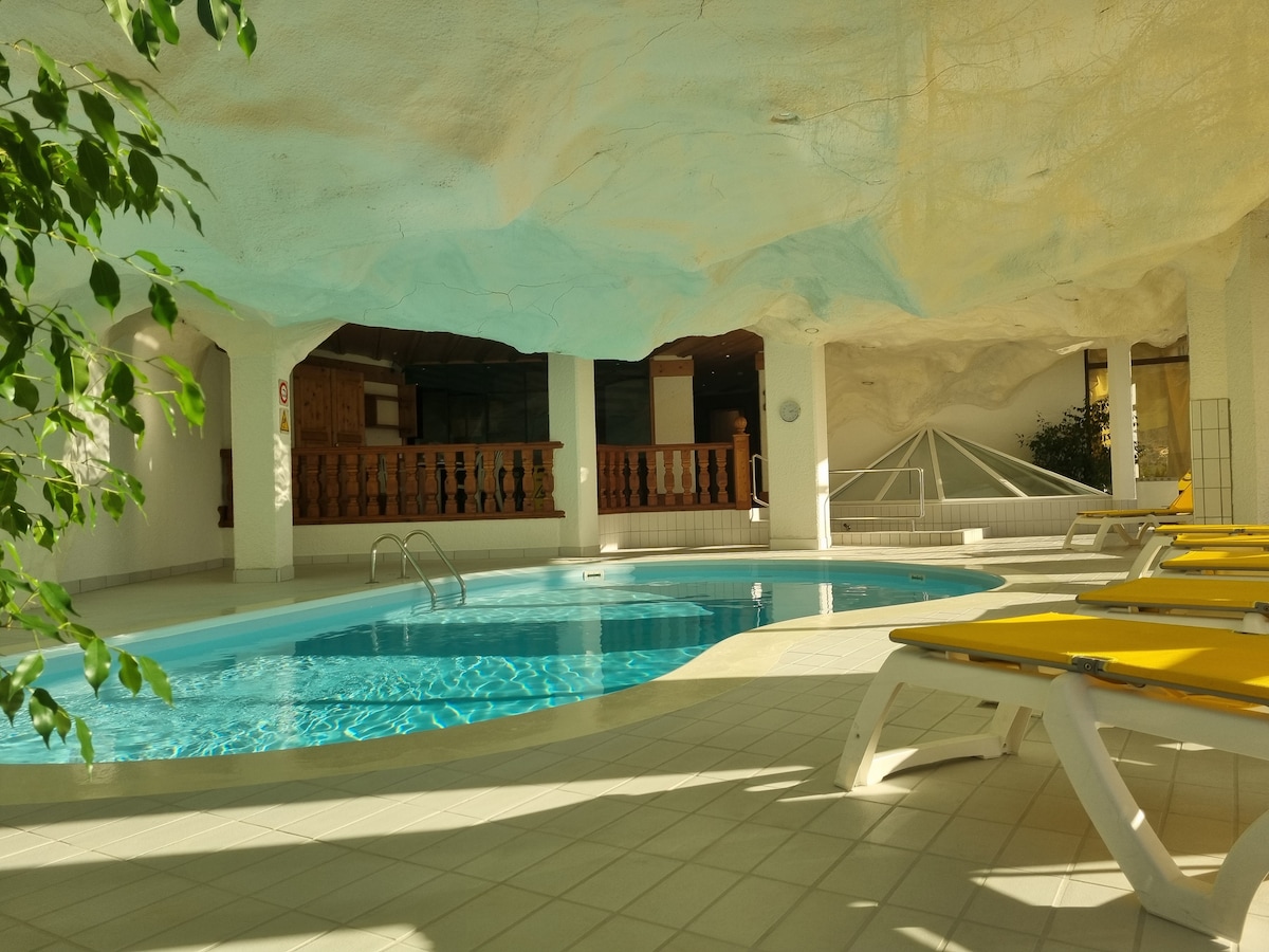 An indoor swimming pool is surrounded by a bright, open space featuring white walls and a unique ceiling design. Sun loungers in cheerful yellow are positioned beside the pool, inviting relaxation. Light filters in from a nearby window, enhancing the tranquil atmosphere.