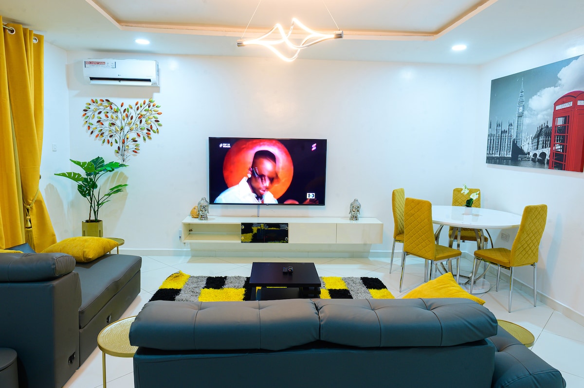 A modern living area is created with a cozy gray couch and vibrant yellow curtains. A flat-screen TV is mounted on the wall, with a stylish coffee table positioned in front. A dining table for four stands nearby, complemented by a decorative plant.
