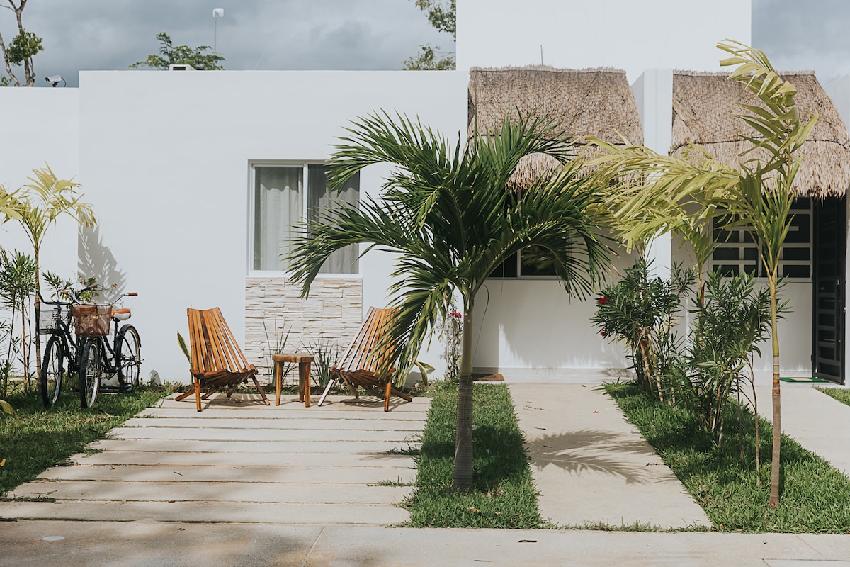 An outdoor area features two wooden lounge chairs positioned on a stone pathway, surrounded by green grass and palm trees. A small table is placed between the chairs. In the background, bicycles are parked, and a thatched roof adds a touch of texture to the setting.