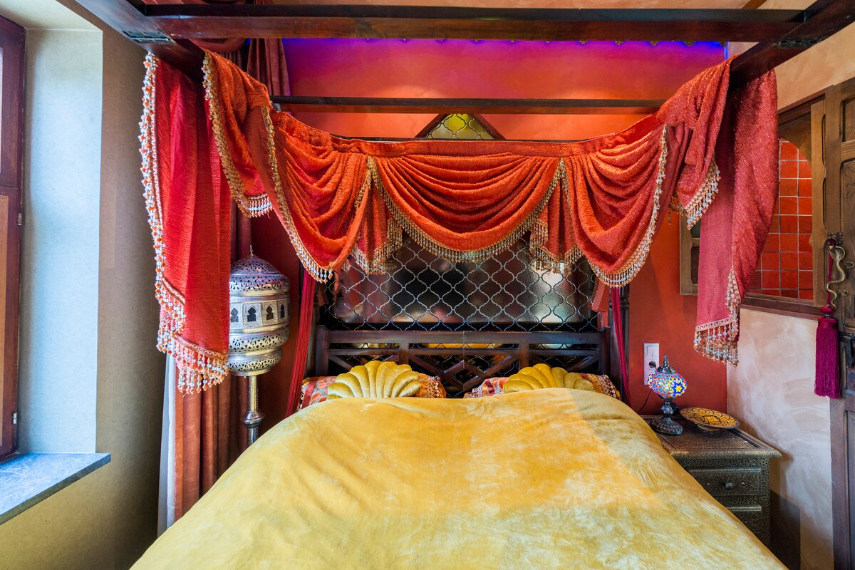 A vibrant sleeping area features a canopied bed draped with rich, flowing fabrics in warm orange hues. Colorful accent lights add a soft glow to the scene. Ornate lamps and intricate detailing enhance the unique character of the room.