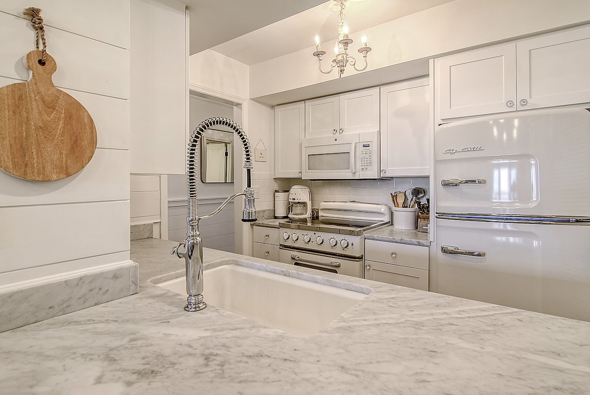 The kitchen features a modern design with marble countertops and white cabinetry. Stainless steel appliances are present, including a gas stove and a refrigerator. A farmhouse sink is showcased, complemented by a stylish pull-down faucet. Natural light is provided through nearby rooms.