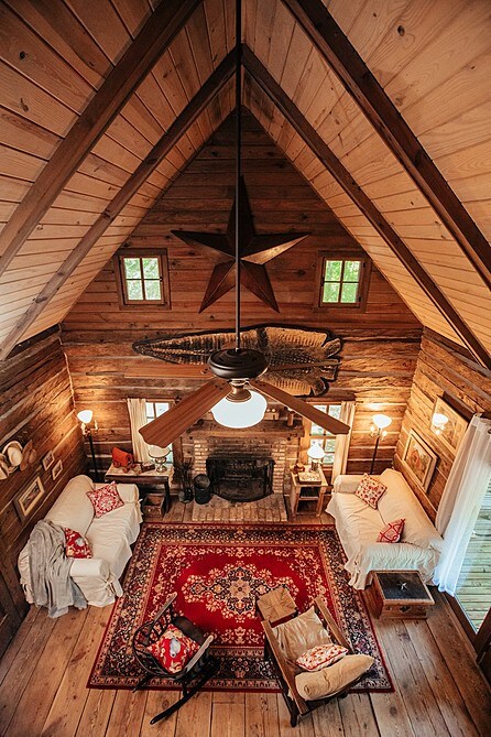 An overview of the living area showcases two sofas across from each other on a rug, with a brick fireplace centered along a wood-paneled wall. Warm lighting emanates from lamps, and wooden beams accentuate the high ceiling. Decorative elements include a star ornament and framed pictures.