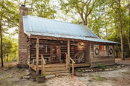 A rustic cabin with a metal roof is framed by surrounding trees. A wooden porch extends at the front, featuring a rocking chair and potted plants. The cabin's weathered wood exterior displays a warm, inviting character, with a brick walkway leading to the entrance.