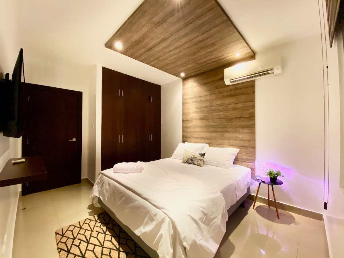 A well-lit bedroom features a spacious bed with crisp white linens and a decorative throw pillow. Natural wood paneling adorns one wall, while a small table with a potted plant adds a touch of greenery. A wall-mounted television and air conditioning unit provide modern amenities.