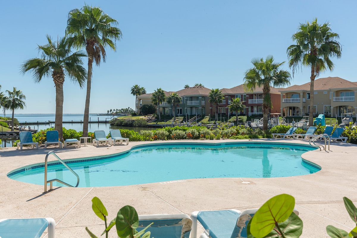 The outdoor pool area is surrounded by lush greenery and palm trees, with comfortable lounge chairs lining the poolside. The clear blue waters of the pool reflect the bright sky, offering a serene view of the nearby waterfront and residential buildings.