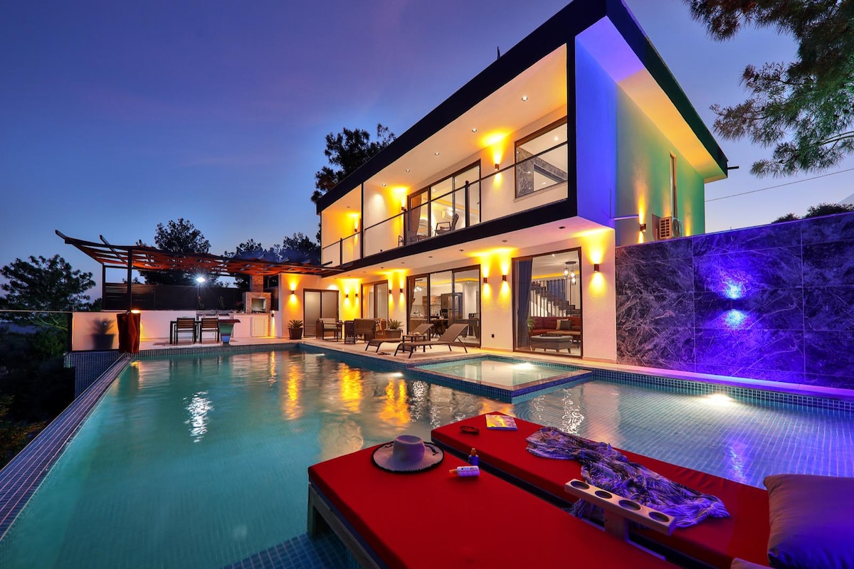 A contemporary villa is illuminated at dusk, revealing a private infinity pool surrounded by sun loungers. The structure features expansive glass doors and a covered outdoor dining area, enhancing the connection to the scenic hillside and distant views.