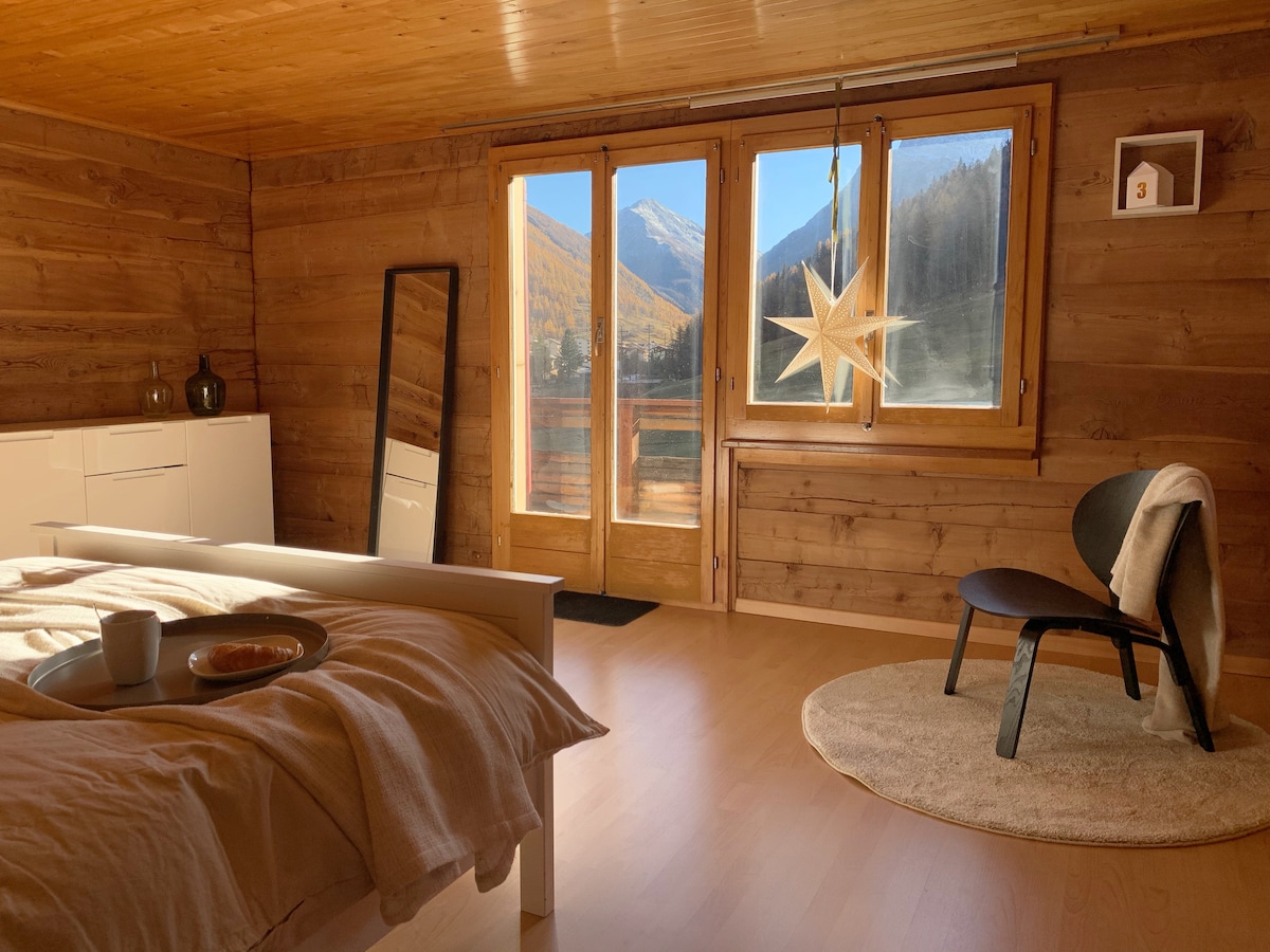 A serene bedroom is designed with wooden paneling and large windows offering views of the mountains. A spacious bed is accompanied by a minimal side table holding a cup and a plate. A decorative star and a cozy chair add charm to the inviting space.